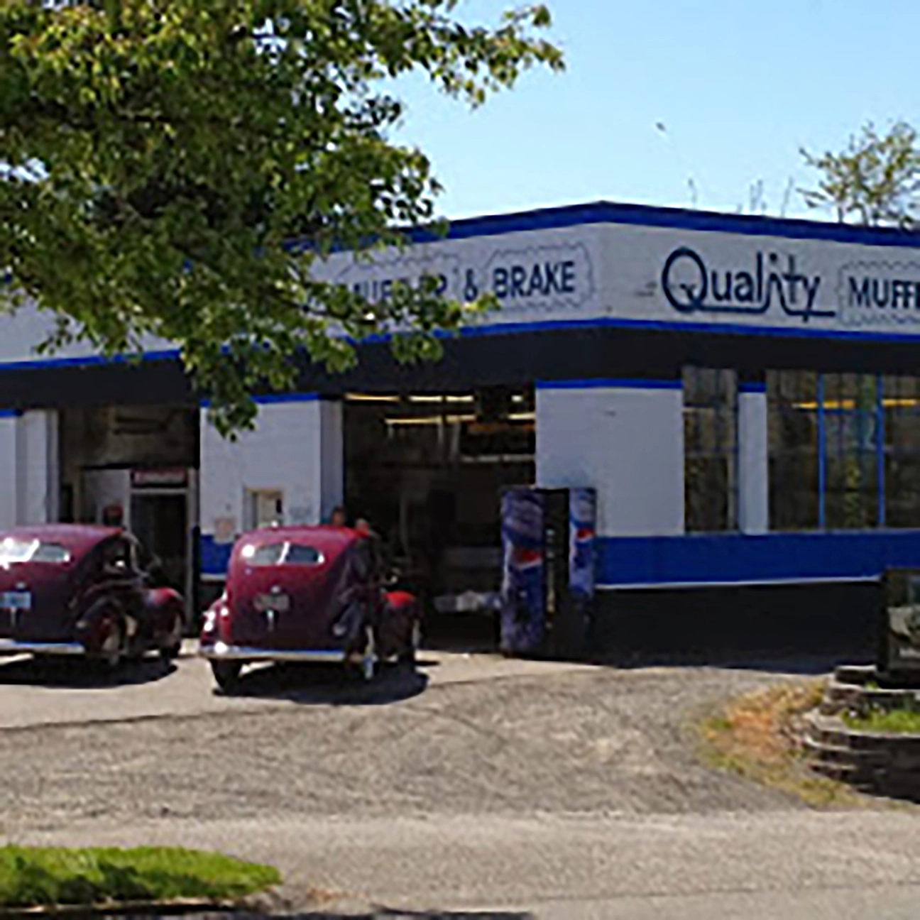 Two vintage dark red cars parked outside the service bays of Quality Muffler and Brake